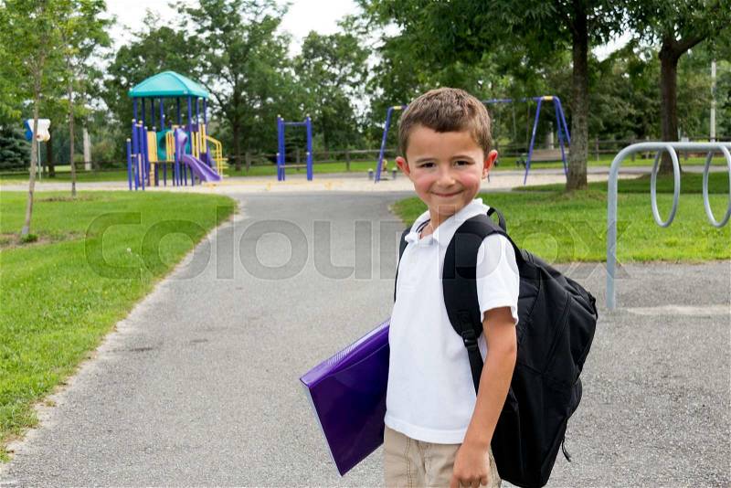Happy little boy with school backpack and book in the play yard, stock photo