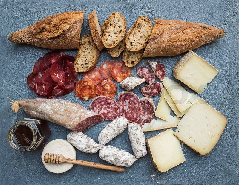 Cheese and meat appetizer selection or wine snack set. Variety of italian cheese, salami, bresaola, baguette and honey on over grey-blue concrete backdrop, top view, stock photo