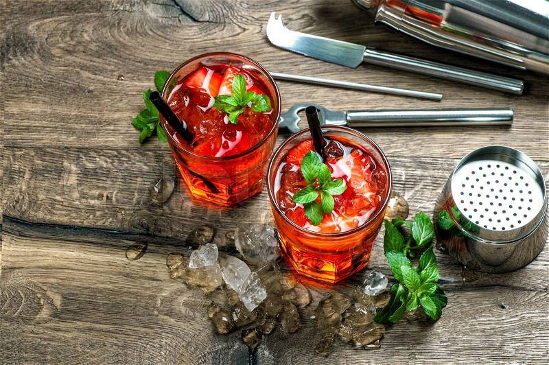 Red cocktail with strawberry, mint leaves, ice. Drink making bar accessories on wooden table, stock photo