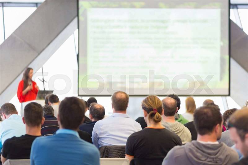 Woman giving presentation in lecture hall. Female speeker having talk at public event. Participants listening to lecture. Rear view, focus on people in audience, stock photo
