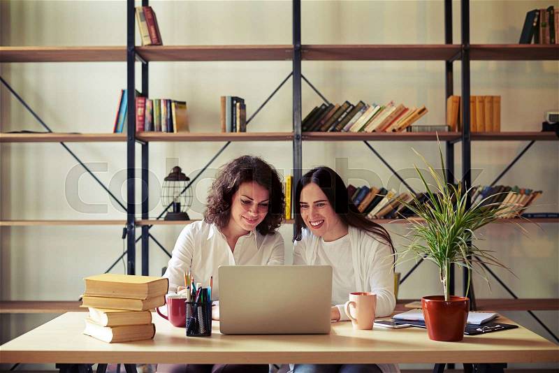 Two women sitting at the table and working at a laptop, bookshelves on a background, stock photo