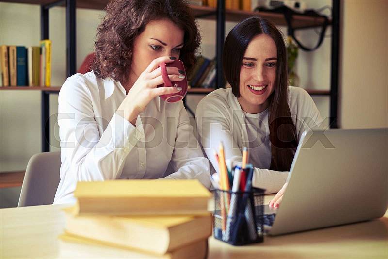 Two women sitting at the table, working at a laptop and drinks, bookshelves on a background, stock photo