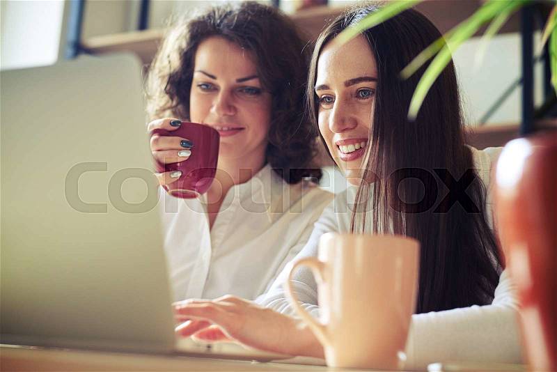 Two women sitting at the table, working at a laptop and drinks, side view, stock photo