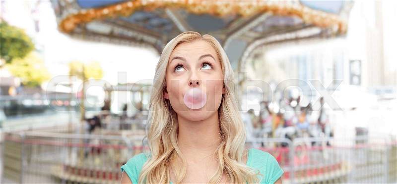 Emotions, expressions and people concept - happy young woman or teenage girl chewing gum over carousel at amusement park background, stock photo