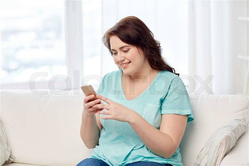People, technology, communication and leisure concept - happy young plus size woman sitting on sofa with smartphone at home, stock photo