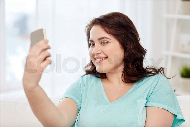 People, technology, communication and leisure concept - happy young plus size woman taking selfie with smartphone at home, stock photo