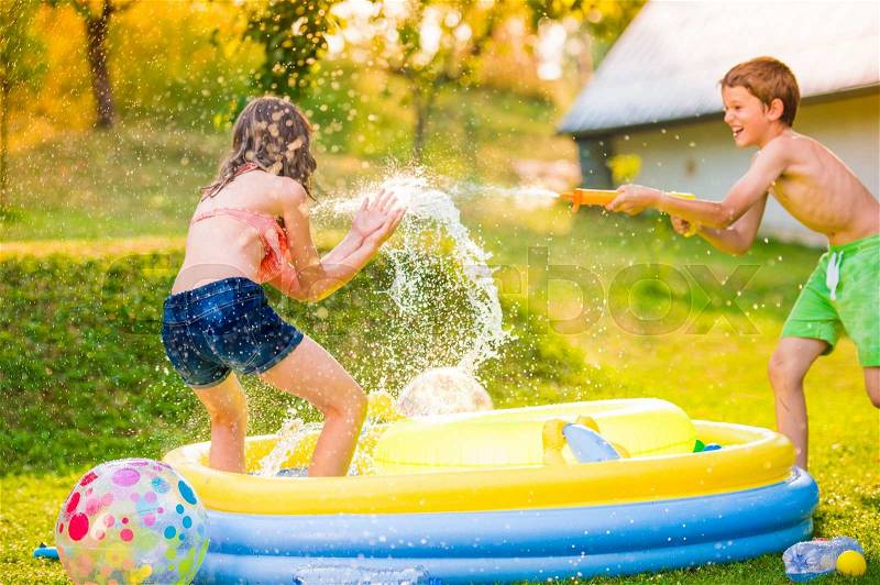 Boy splashing girl with water gun in garden swimming pool, sunny summer day, back yard, stock photo