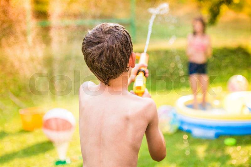 Boy splashing girl with water gun in garden swimming pool, sunny summer day, back yard, rear view, stock photo