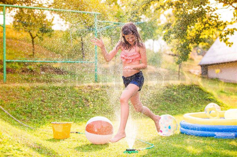 Girl running above a sprinkler, sunny summer back yard, garden swimming pool, stock photo