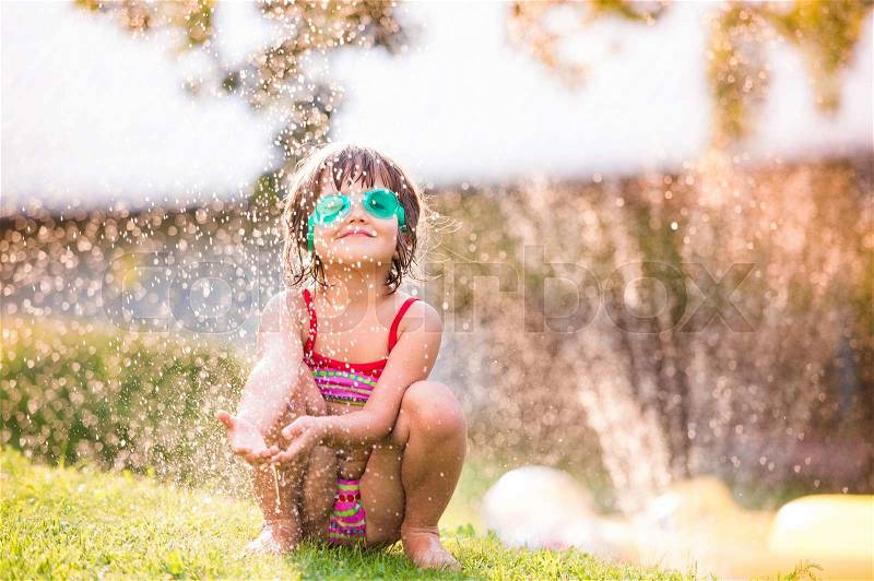 Cute girl crouching in the garden under the water splashing from sprinkler, sunny summer back yard, stock photo