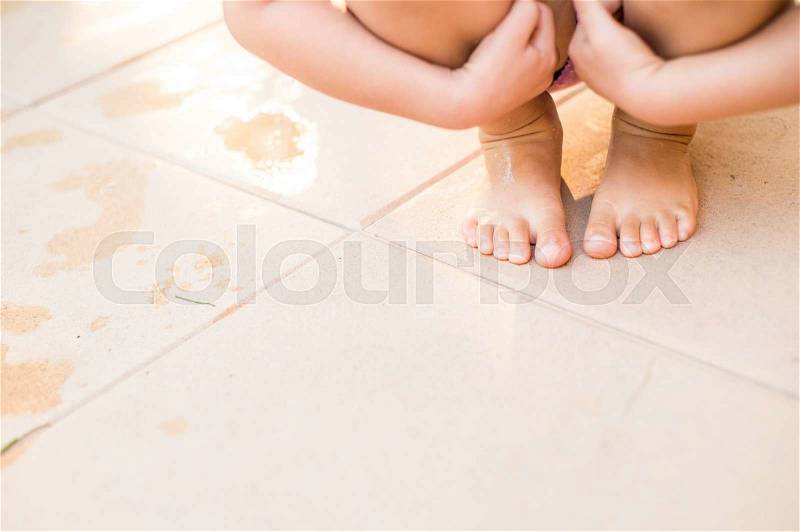 Unrecognizable child in swimsuit crouching on pavement, holding her knees, wet footprints, copy space, stock photo