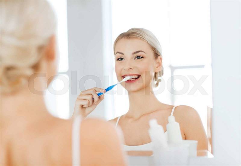 Health care, dental hygiene, people and beauty concept - smiling young woman with toothbrush cleaning teeth and looking to mirror at home bathroom, stock photo
