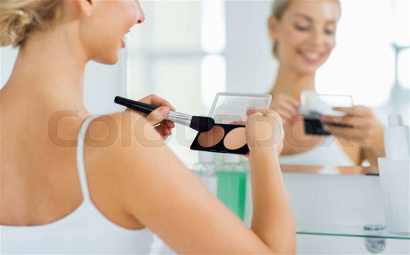 Beauty, make up, cosmetics, morning and people concept - close up of happy young woman with makeup brush and contouring and highlighting palette looking to mirror at home bathroom, stock photo