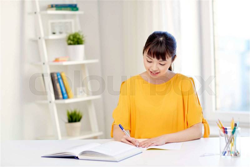 People, education, high school and learning concept - happy asian young woman student with book and notepad writing at home, stock photo