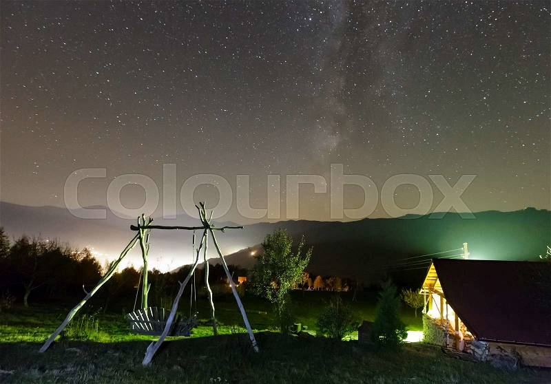 Milky Way galaxy in starry night sky and rural yard illuminated in green color on mountain hill, stock photo