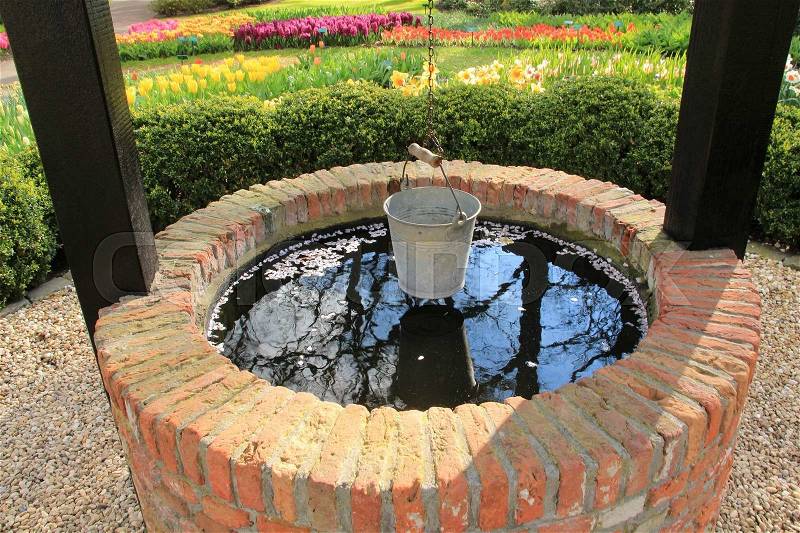 At the foreground a wishing well with a hanging bucket and at the background a bed of blooming tulips and daffodils in the flower garden, Keukenhof, in Lisse in spring, stock photo
