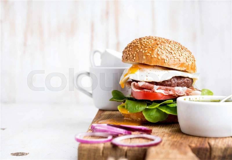 Breakfast set. Homemade beef burger with fried egg and vegetables, onion rings and coffee cups on wooden board, white painted background. Selective focus, copy space, stock photo