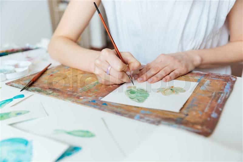 Closeup of hands of woman artist painting with paintbrush and watercolor paints on the table , stock photo