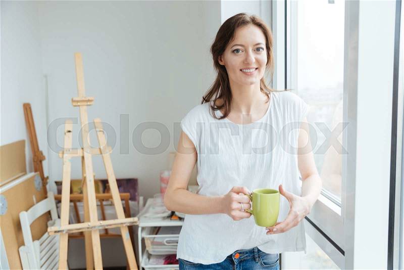 Cheerful beautiful woman painter drinking tea near the window in art studio, stock photo