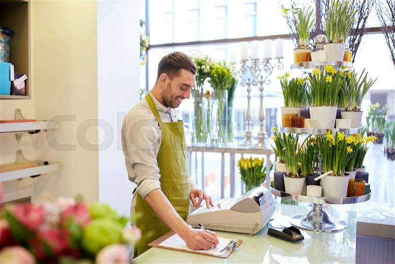 People, sale, retail, business and floristry concept - happy smiling florist man with clipboard and cashbox writing and making notes order at flower shop counter, stock photo