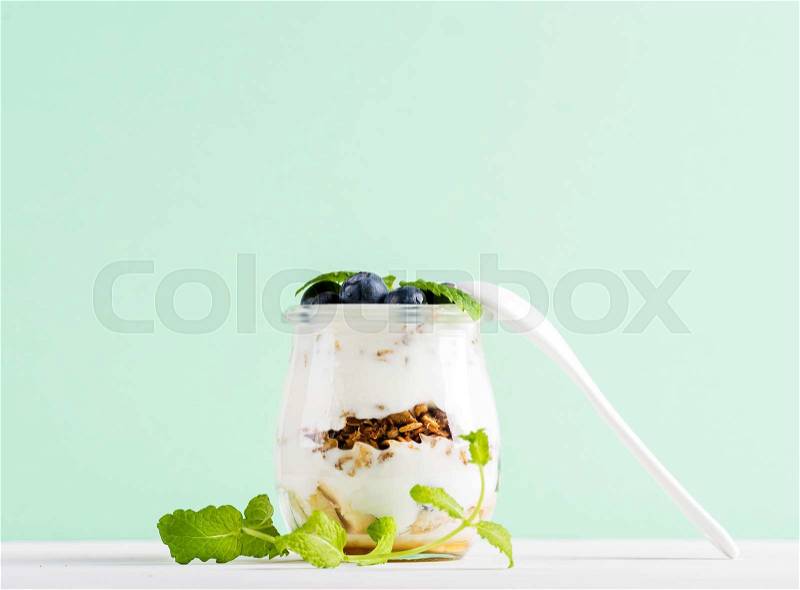 Yogurt oat granola with jam, blueberries and green leaves in glass jar on mint pastel backdrop, stock photo