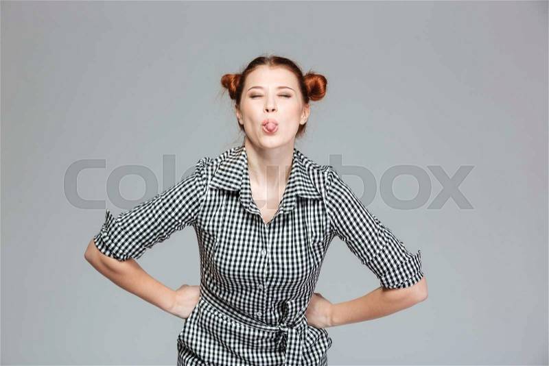 Amusing cute young woman showing tongue and making funny face over grey background , stock photo
