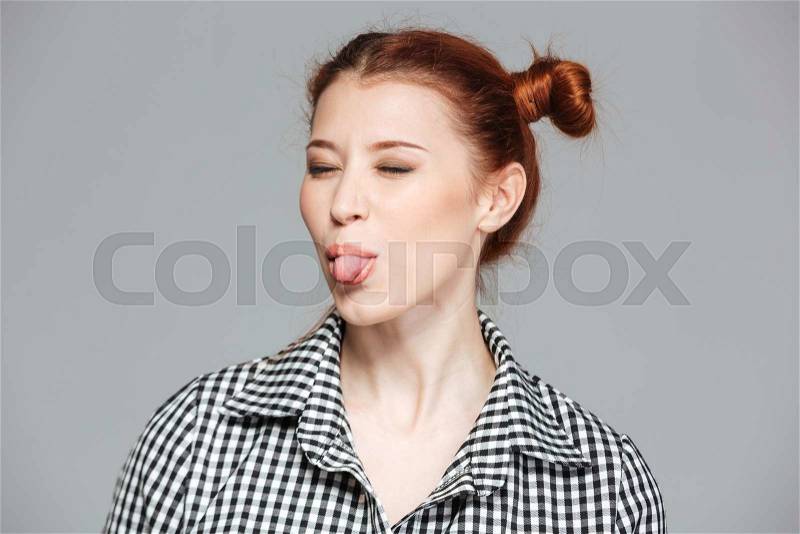 Funny pretty young woman with eyes closed showing tongue over grey background, stock photo