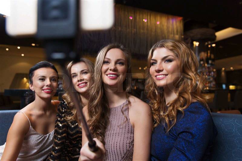 Celebration, friends, bachelorette party, technology and holidays concept - happy women with smartphone selfie stick taking picture at night club, stock photo