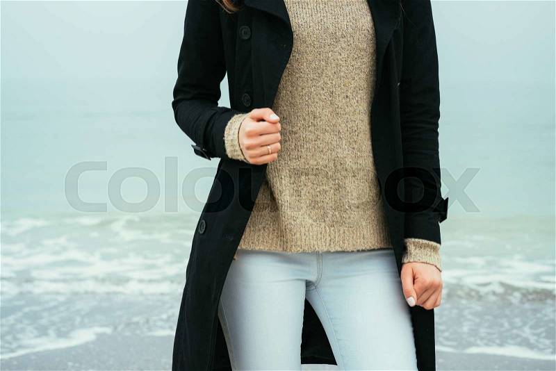 Slim woman in jeans, a sweater and a black coat against the sea on a cloudy day, stock photo