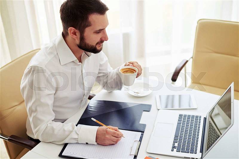 Beard man sitting on a comfortable chair writes something in a pad and drink a coffee in the white modern office, stock photo