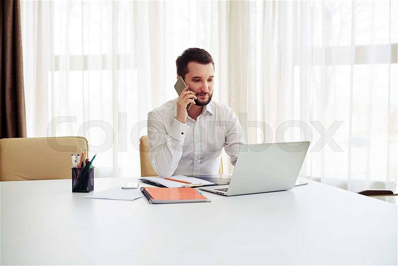 Man working on the laptop and calling someone on the phone in the modern white office, stock photo