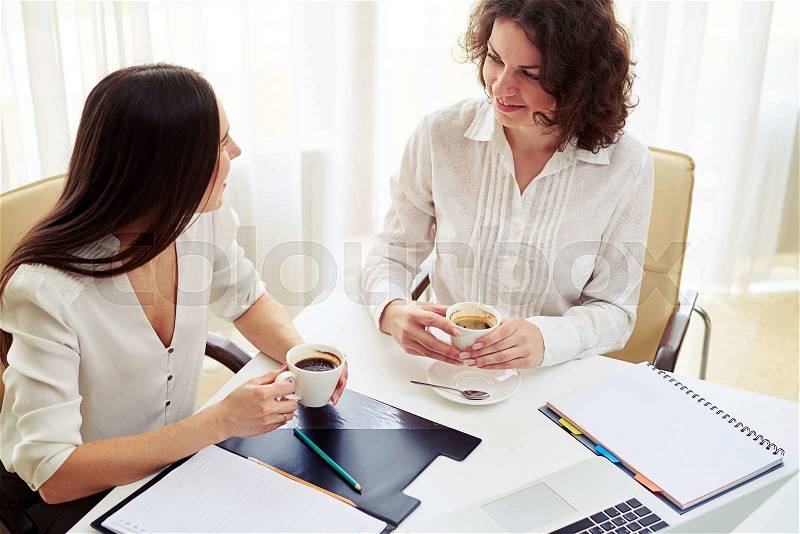 Two young beautiful women talking about something and drink coffee in the spacious office, stock photo