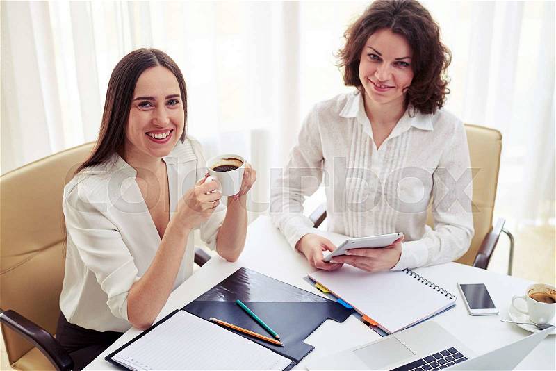 Two young smiling women teamworking with gadgets and drinking coffee in the modern office, stock photo