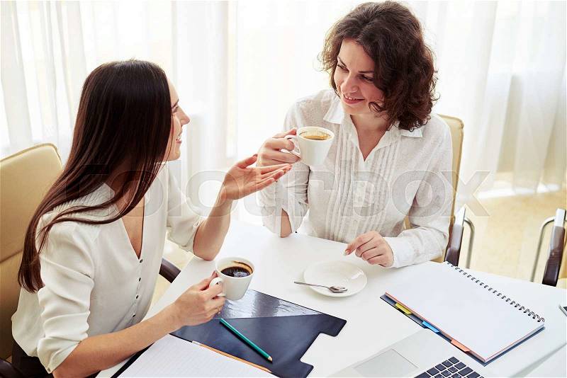 Two young pretty women talking about something and hold coffee in hand in the spacious office, stock photo
