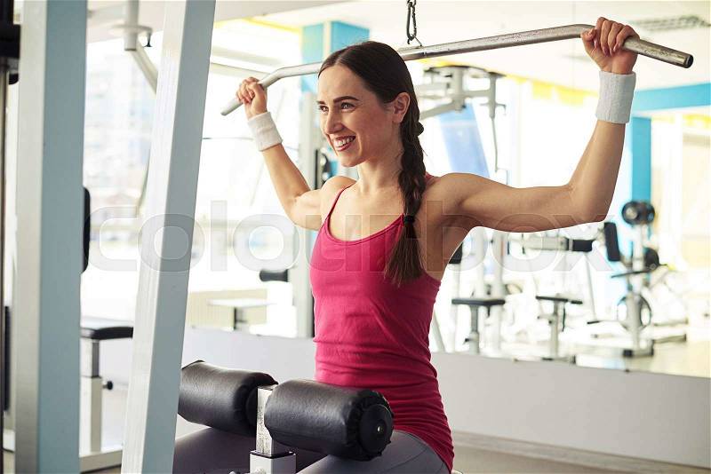Young beautiful smiling woman is working out pull-down machine in gym near mirror, stock photo