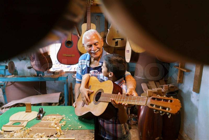 Small family business and traditions: old grandpa with grandson in lute maker shop. The senior artisan gives teaches how to play classic guitar to the boy, who plays his first notes with the instrument, stock photo