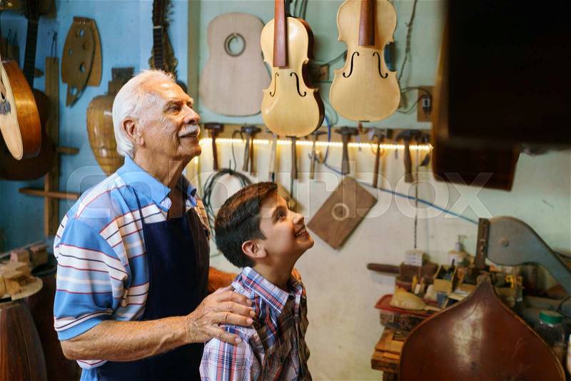 Small family business and traditions: old grandpa with grandson in lute maker shop. The senior artisan hugs the boy and shows him his handmade guitar and music instruments, stock photo