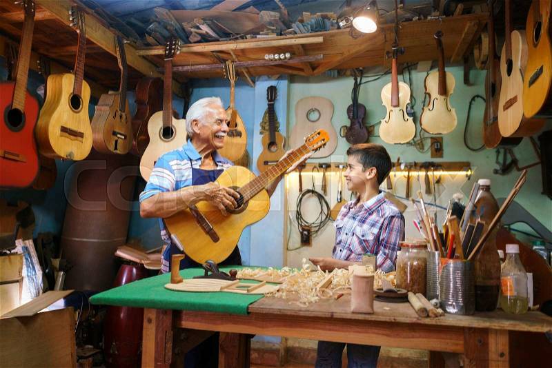 Small family business and traditions: old grandpa with grandson in lute maker shop. The senior artisan gives teaches how to play classic guitar to the boy, who looks carefully at the instrument, stock photo