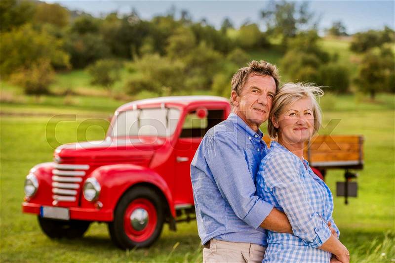 Senior couple hugging, vintage styled red car, green sunny nature, stock photo