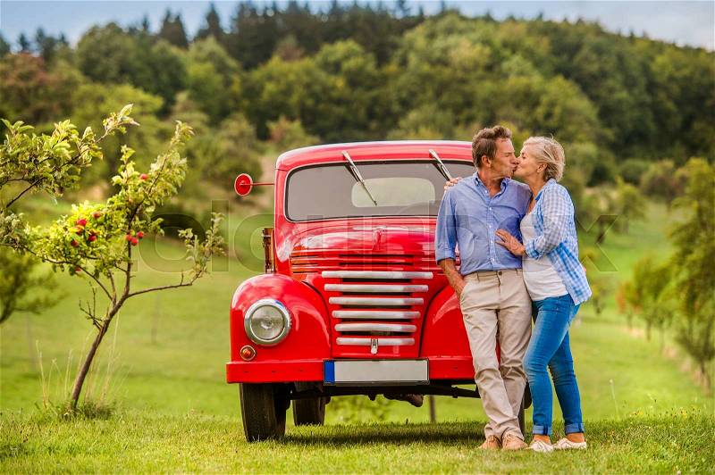 Senior couple hugging and kissing, vintage styled red car, green sunny nature, stock photo