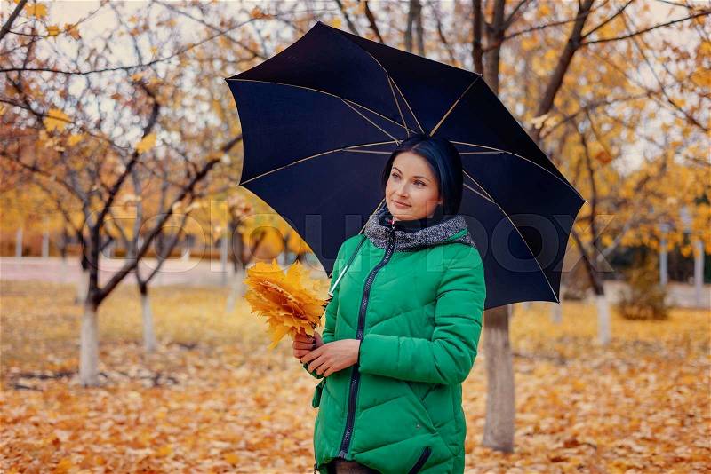 Cute young smiling woman in green winter coat holding large yellow maple tree leaves and umbrella outdoors in autumn scene, stock photo