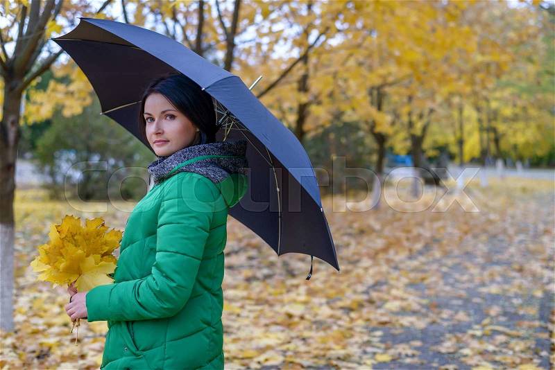 Calm single woman standing under umbrella on sidewalk covered with leaves along path of maple trees in autumn theme portrait, stock photo