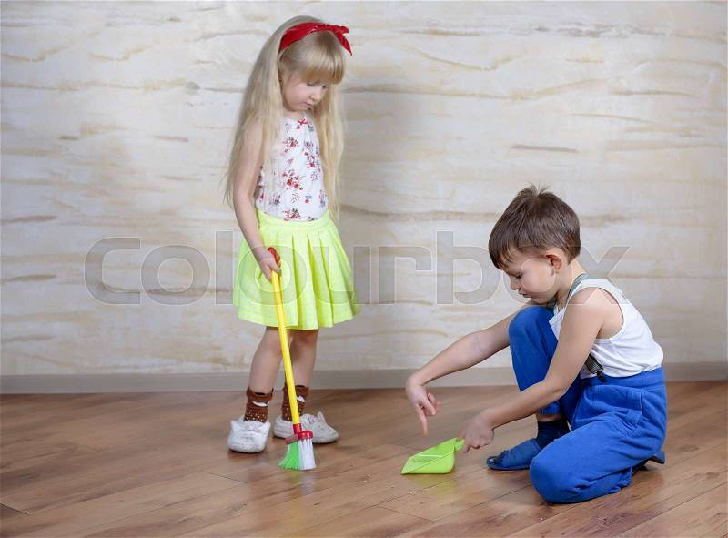 Cute little boy in blue pants, slippers and suspenders using toy broom and dustpan with girl in green skirt on hardwood floor, stock photo