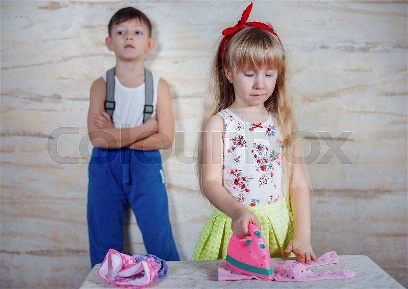 Serious little boy in blue pants held up by suspenders standing with folded arms against wall while girl irons pink shirt, stock photo