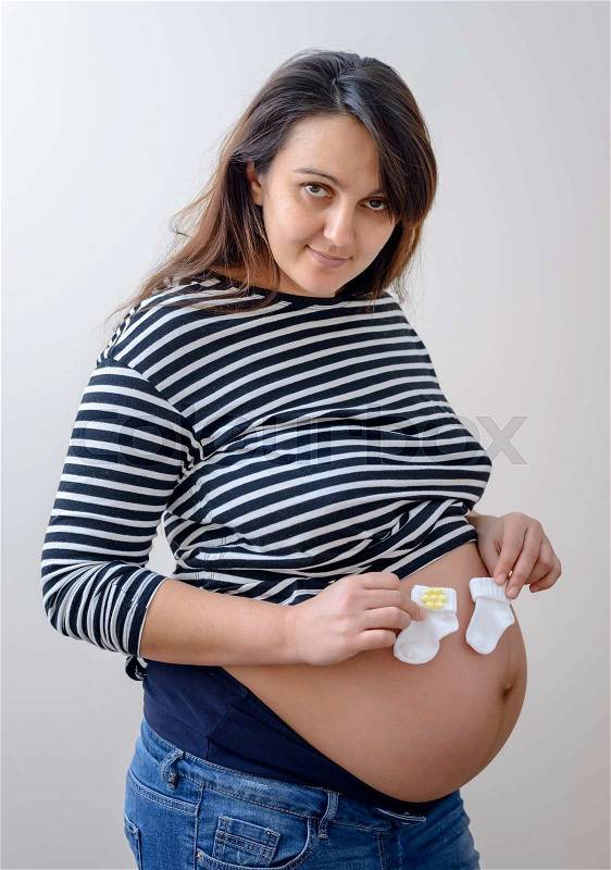 Pregnant woman holding tiny white baby booties over her heavily pregnant stomach with a smile of anticipation, stock photo