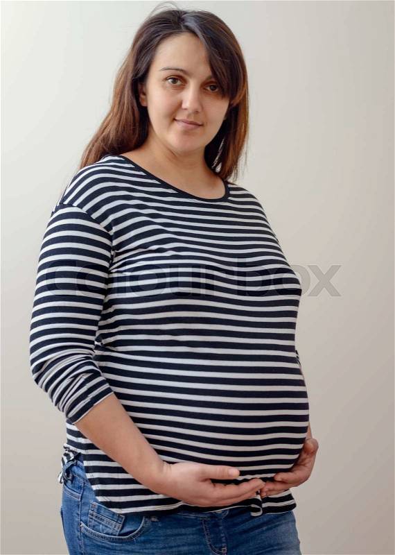 Cute expecting woman in striped shirt holding her protruding large belly over gray background, stock photo
