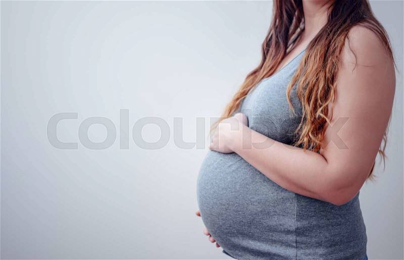 Close up on unidentifiable single pretty pregnant woman in gray shirt holding belly while in front of gray background with copy space, stock photo
