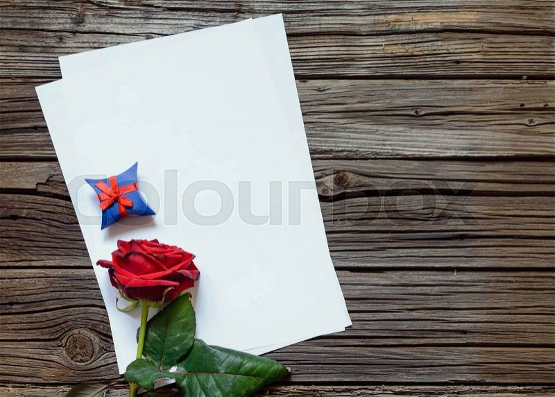 Pair of stacked blank papers centered on table with red rose and little blue gift box, stock photo