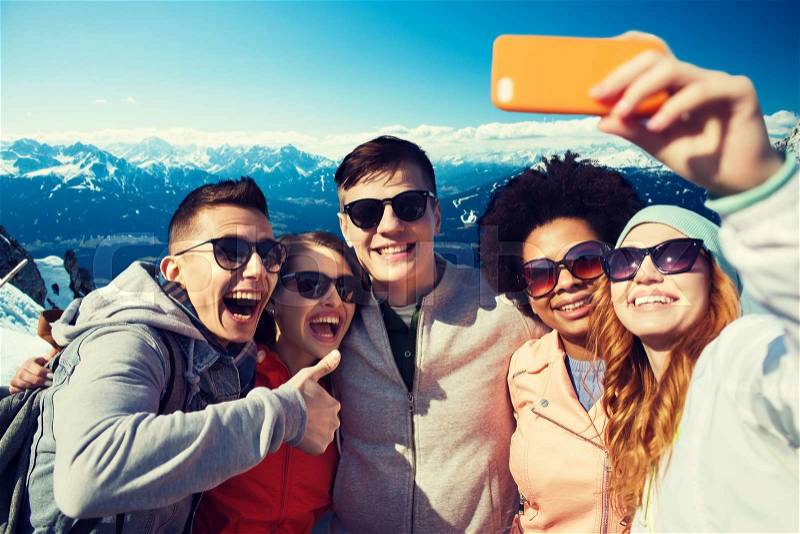 People, travel, tourism, friendship and technology concept - group of happy teenage friends taking selfie with smartphone and showing thumbs up over alps mountains in austria background, stock photo