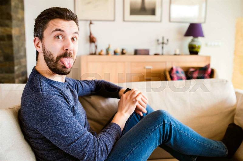 Man at home sitting on sofa using smart watch, making funny face, sticking tongue out, stock photo